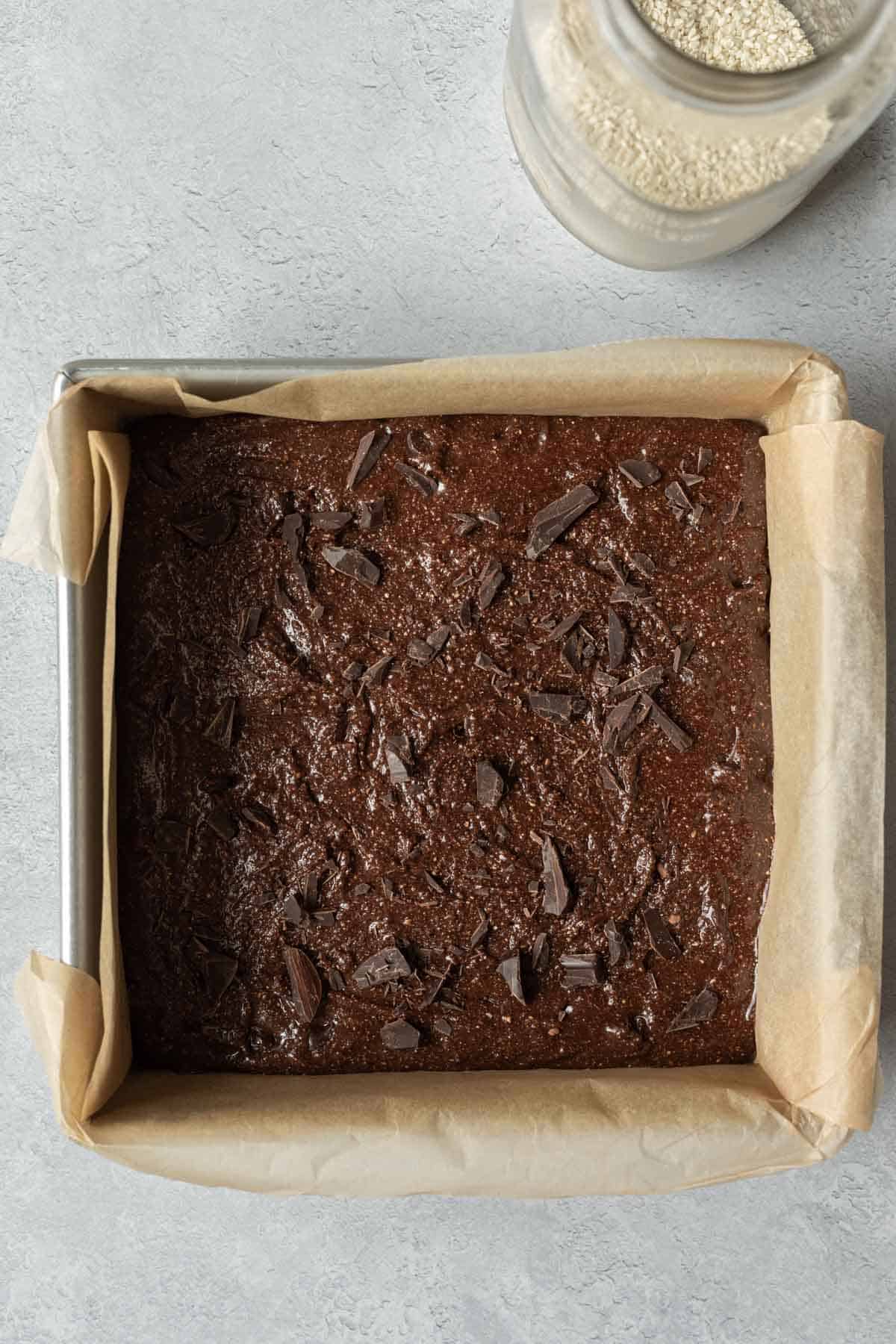 Brownie batter in parchment-lined pan with dark chocolate shards sprinkled on top.