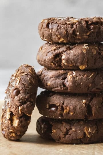 A stack of 5 chocolate protein cookies with one cookie leaning against.