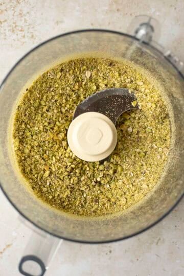 Inside the bowl of a food processor with ground pistachios and oats.