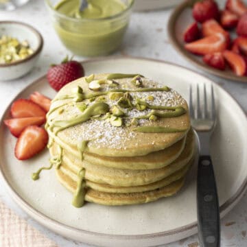 A stack of five pistachio pancakes on a plate topped with pistachio cream and served with fresh strawberries.