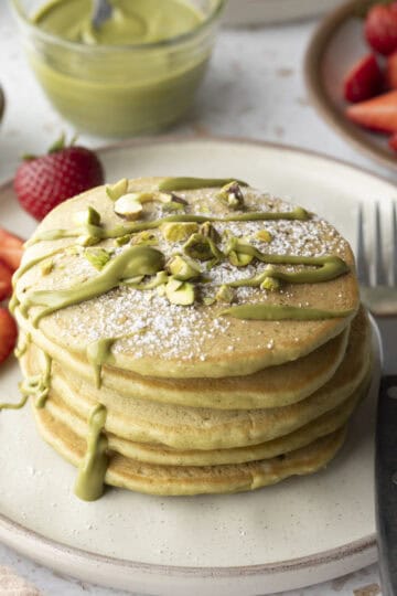 A stack of five pistachio pancakes on a plate topped with pistachio cream and served with fresh strawberries.