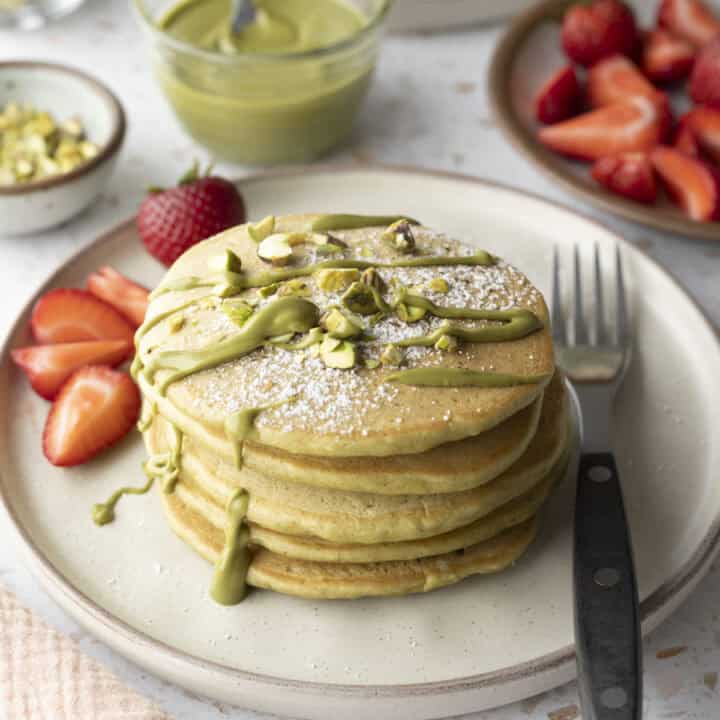 A stack of five pistachio pancakes on a plate topped with pistachio cream and served with fresh strawberries.