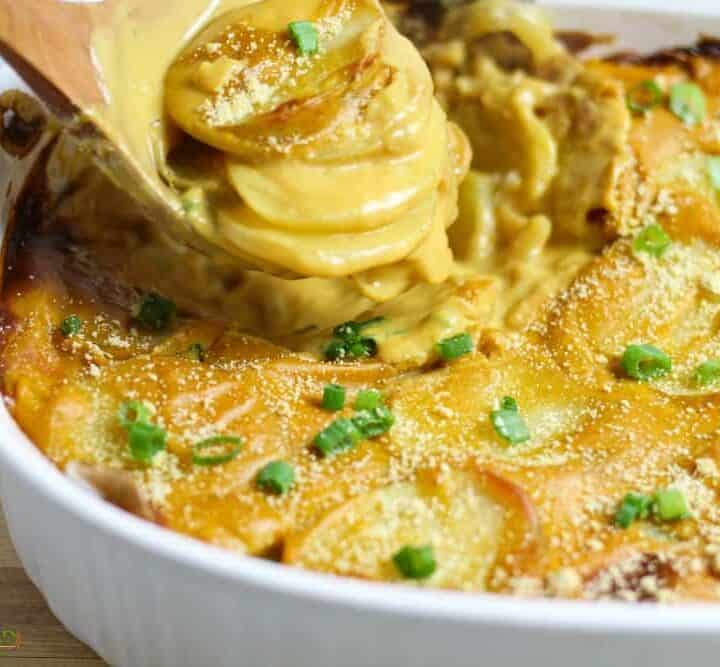A spoon scooping up cheesy vegan scalloped potatoes from a white baking dish.