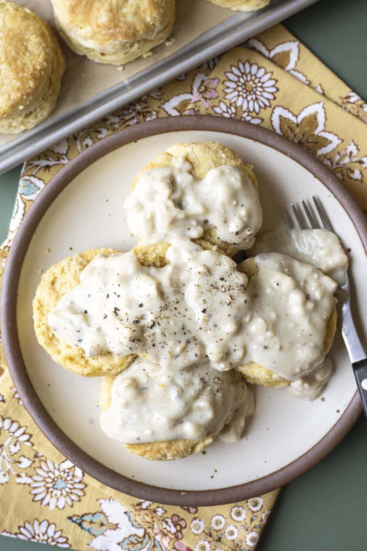 Biscuits on a plate covered with vegan sausage gravy with more biscuits on a baking sheet.