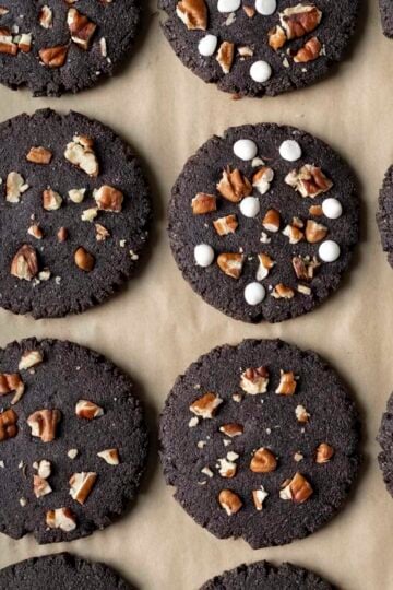 Black cocoa cookies topped with white chocolate chips and chopped pecans resting on parchment paper.