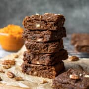 A stack of five pumpkin brownies with chopped pecans and a bowl of pumpkin puree nearby.