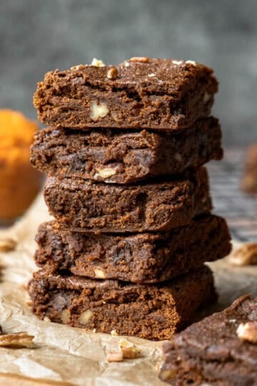 A stack of five pumpkin brownies with chopped pecans and a bowl of pumpkin puree nearby.