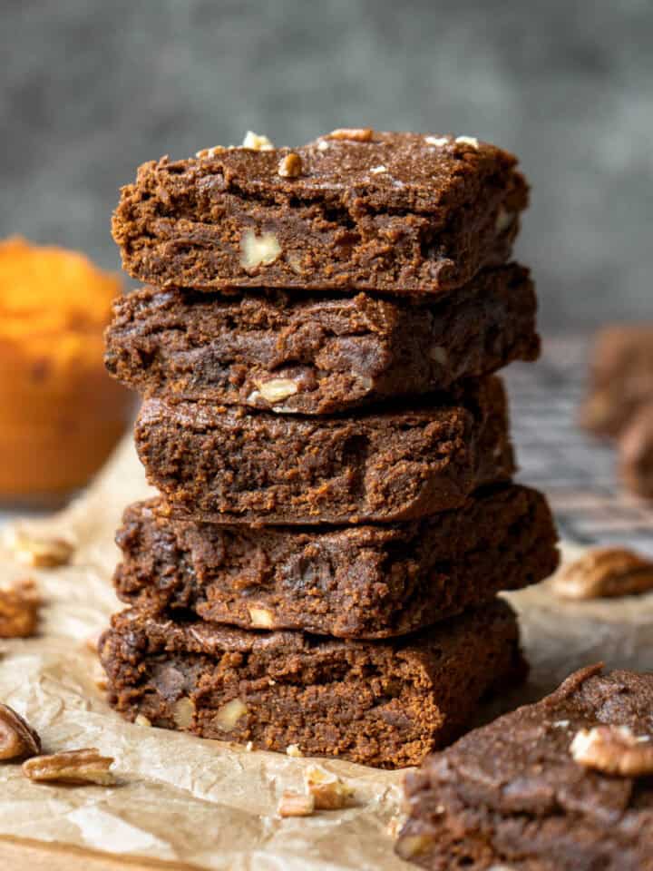 A stack of five pumpkin brownies with chopped pecans and a bowl of pumpkin puree nearby.