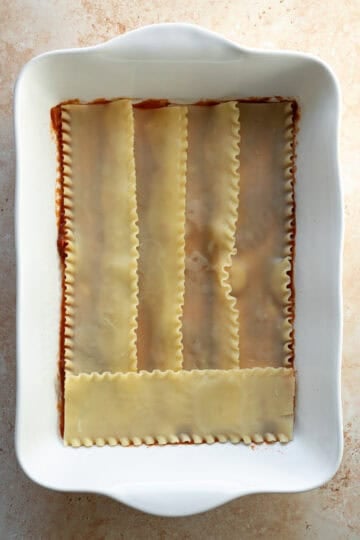 A thin layer of marinara sauce in a large baking dish with 5 lasagne noodles laid on top.