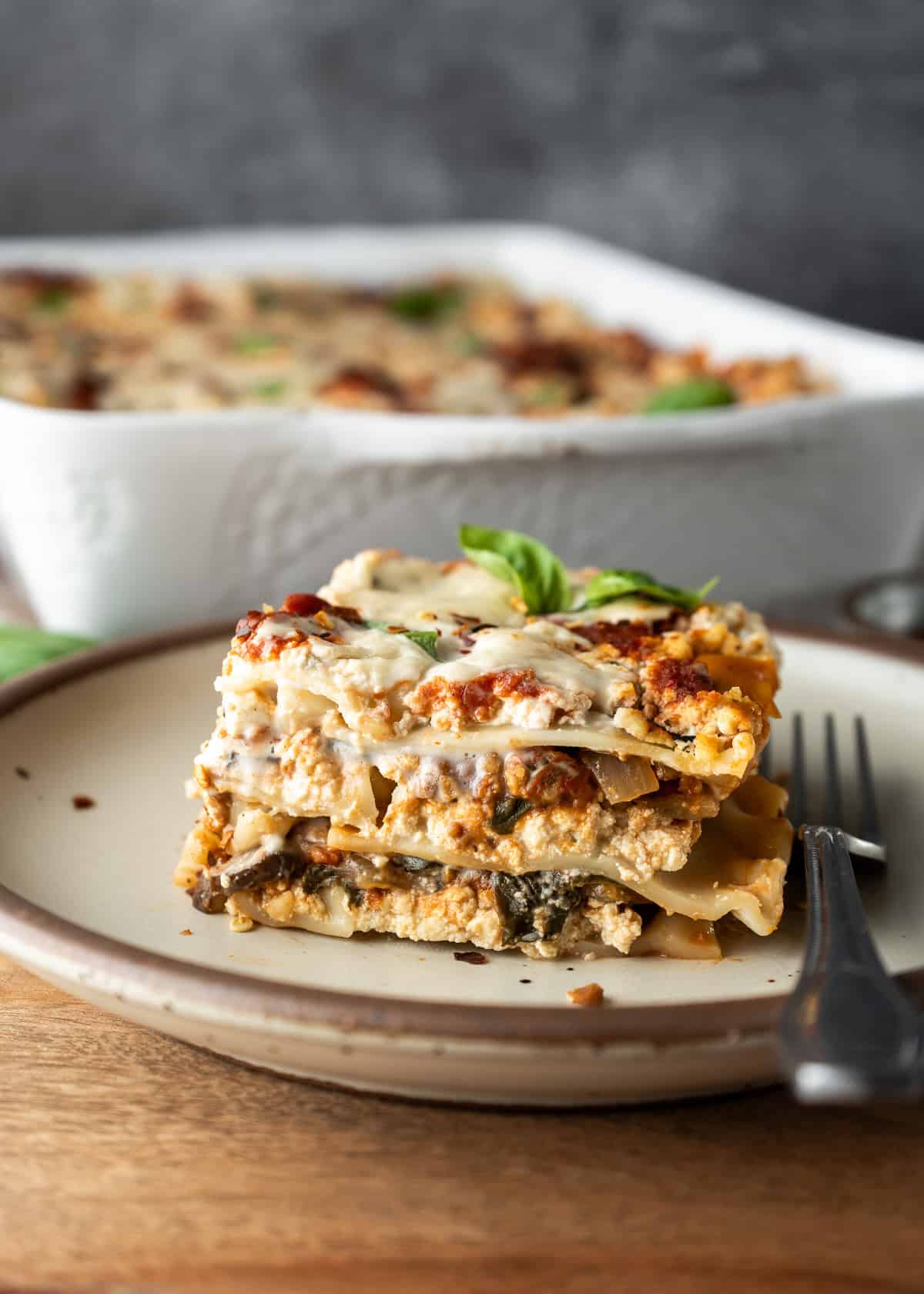 A slice of veggie lasagna on a plate with a fork resting nearby and the lasagna dish in background.