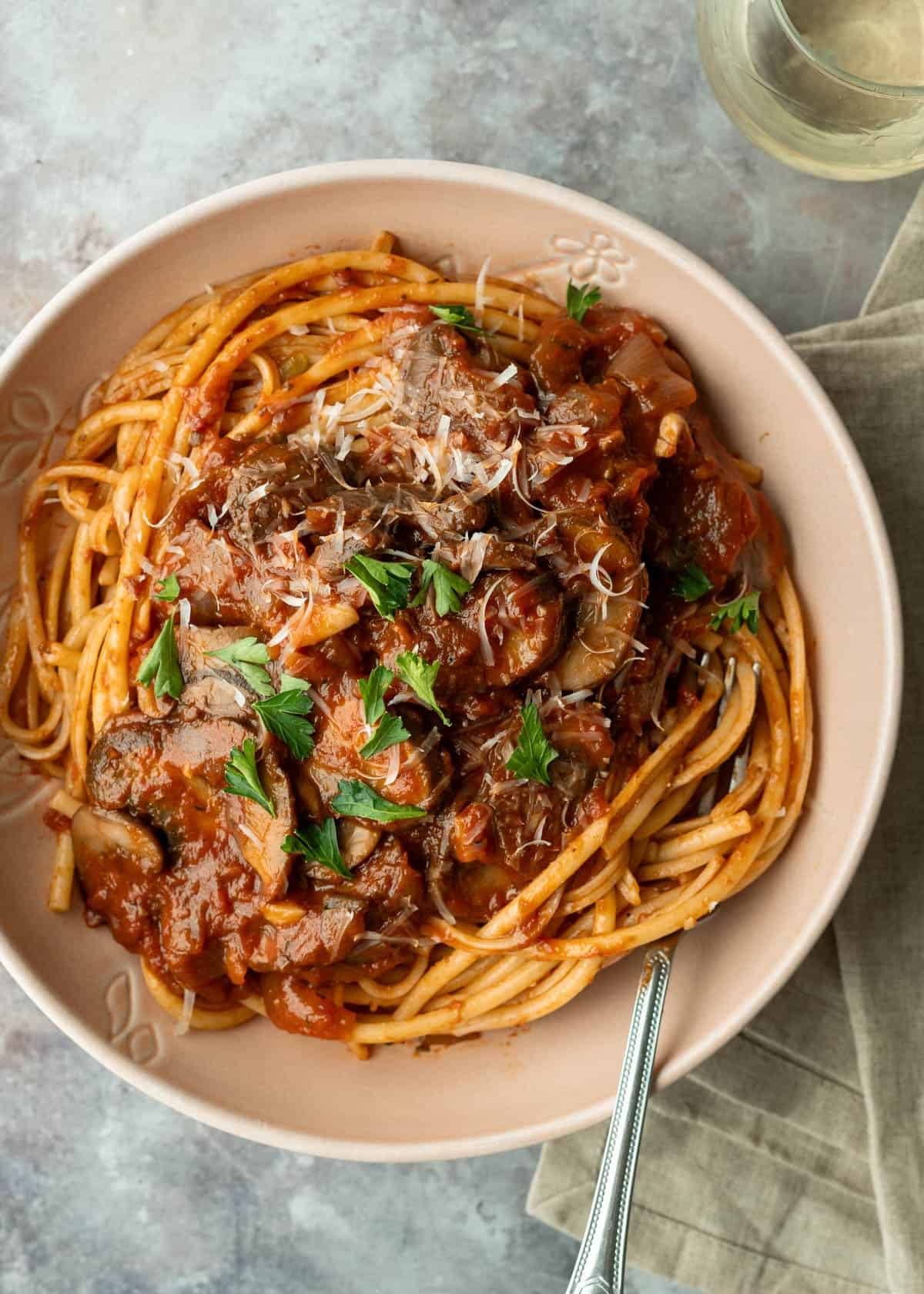 Rich mushroom ragu over bucatini pasta with a glass of wine nearby.