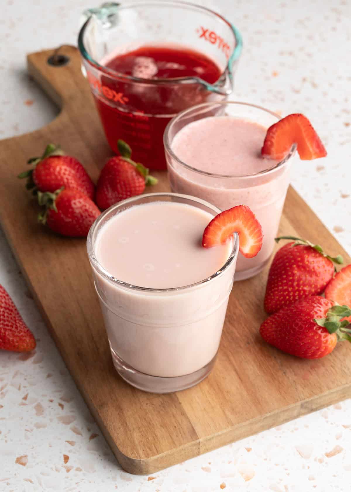 Two glasses of homemade strawberry oat milk with a cup of strawberry syrup in the background.