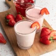 Two glasses of strawberry milk on a wood board with fresh strawberries nearby.