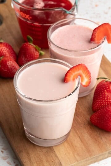 Two glasses of strawberry milk on a wood board with fresh strawberries nearby.