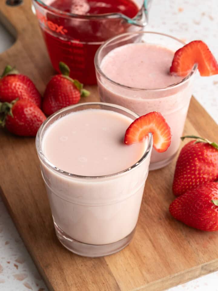 Two glasses of strawberry milk on a wood board with fresh strawberries nearby.