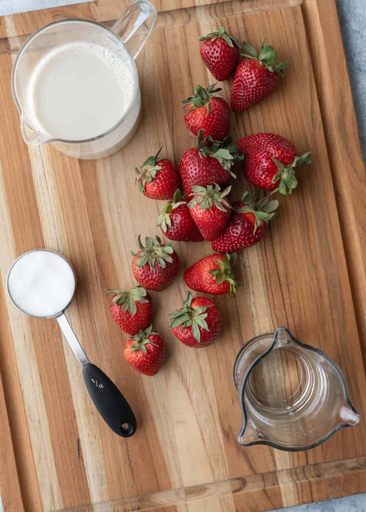 Fresh strawberries, oat milk, sugar, and water on a wood board.