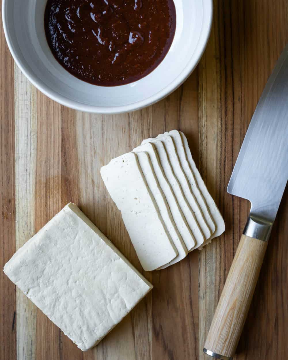 Block of tofu being sliced and a bowl of thick marinade sitting nearby.