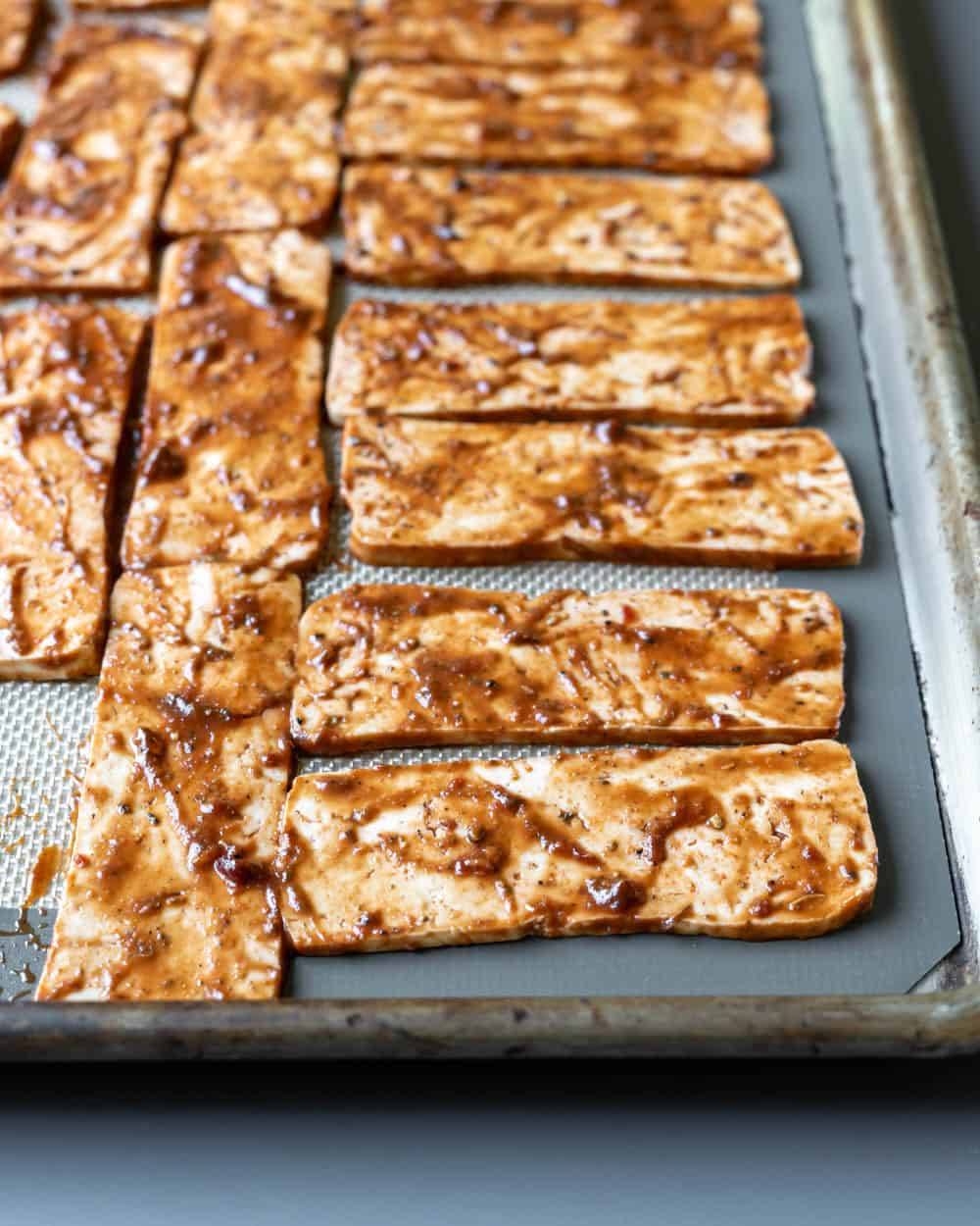 Thin slices of tofu coated in spicy thick marinade spread out on a baking sheet.