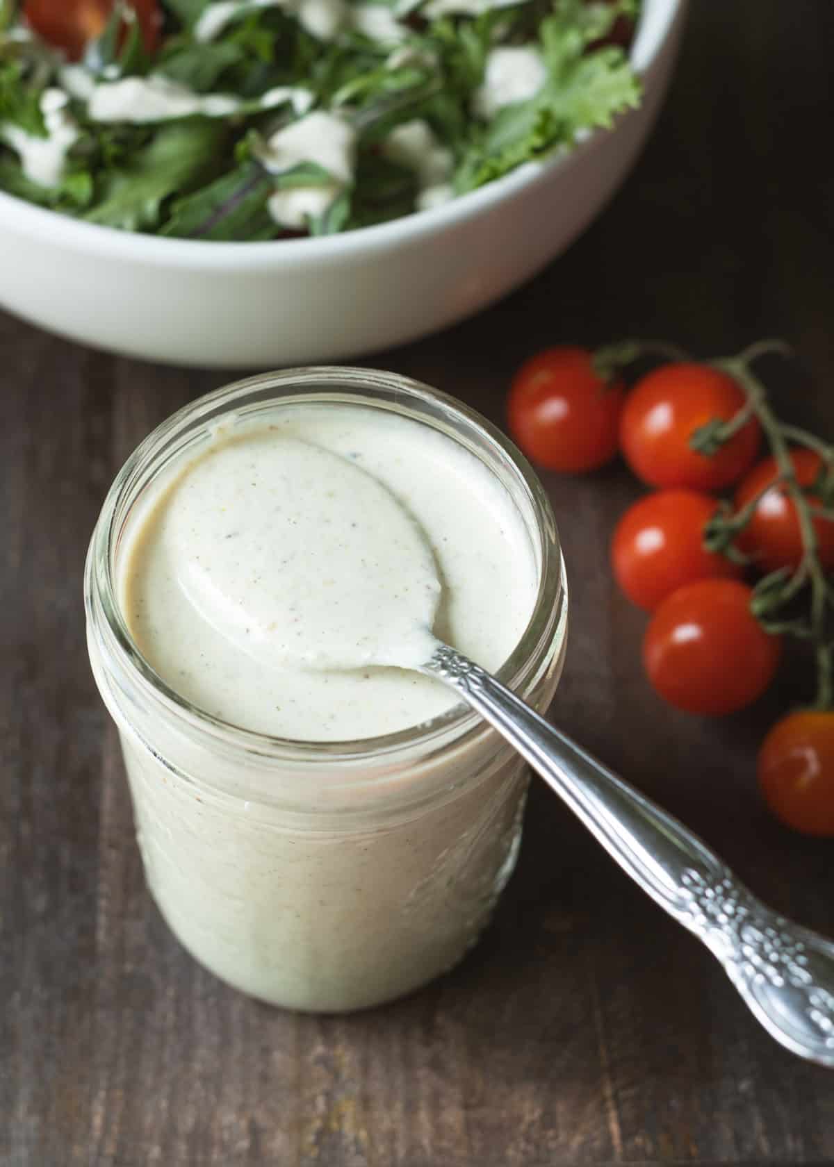 Creamy pepperoncini dressing in a glass jar with salad and tomatoes in background.