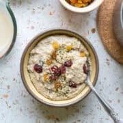 A spoon resting in a bowl of fonio porridge topped with seeds and dried fruit.