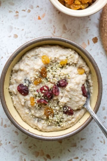 A spoon resting in a bowl of fonio porridge topped with seeds and dried fruit.