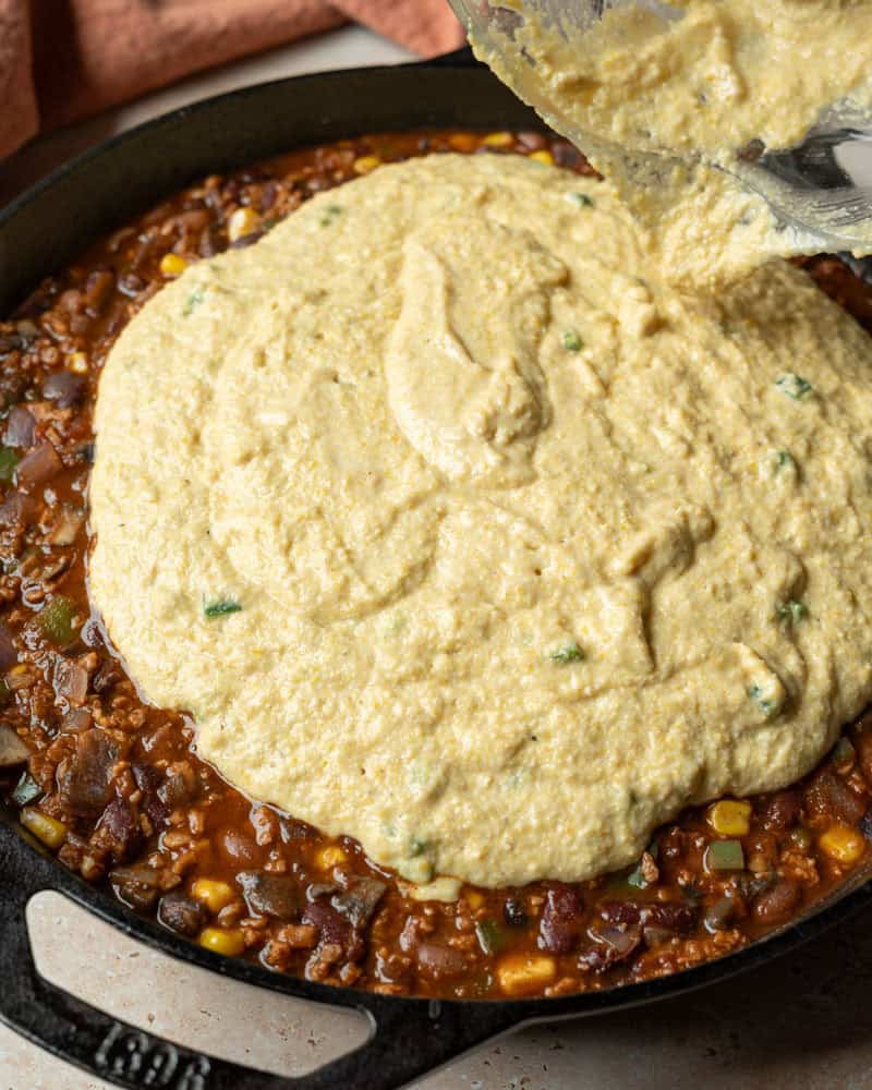 Pouring cornbread batter over thick chili in a skillet.