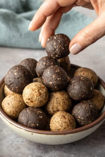 Two flavors of homemade protein balls piled in a bowl with a hand picking up one.
