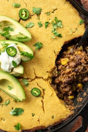 Vegan chili cornbread casserole in a cast iron skillet topped with avocado and cilantro.
